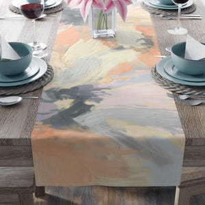 May include: A table setting with a colorful abstract art table runner. The runner features a blend of peach, gray, and cream hues. The table is set with teal-colored bowls, plates, and wine glasses. A vase of pink lilies sits in the center.