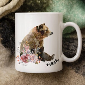 Może przedstawiać: Biały kubek ceramiczny z ilustracją brązowego niedźwiedzia otoczonego różowymi i czerwonymi kwiatami. Na kubku nadrukowany jest napis "Sarah".