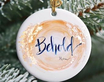 May include: A white ceramic Christmas bauble with a gold and peach watercolour background. The bauble features the words "Behold Him" in blue calligraphy.