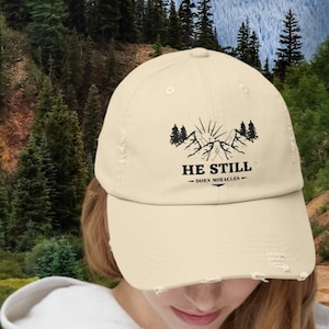Op de afbeelding: Een beige, verweerde baseballpet met een zwart berglandschap en de tekst "HE STILL DOES MIRACLES". De pet wordt gedragen door een persoon die voor een bergketen en een rivier staat.