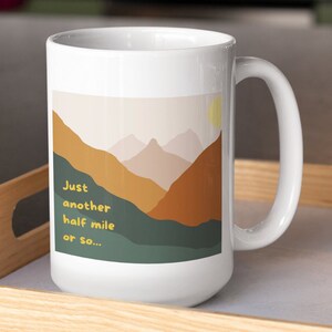 May include: White ceramic mug with a mountain landscape design. The mug has a handle and says "Just another half mile or so..." in yellow text.