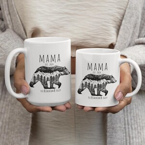 May include: Two white ceramic mugs with black handles. Both mugs feature a black and white illustration of a bear with a mountain range and trees inside its silhouette. The text "MAMA EST. 2024" is printed on both mugs. The text "ELL JACKSON & LILLY" is printed on the bottom of both mugs.