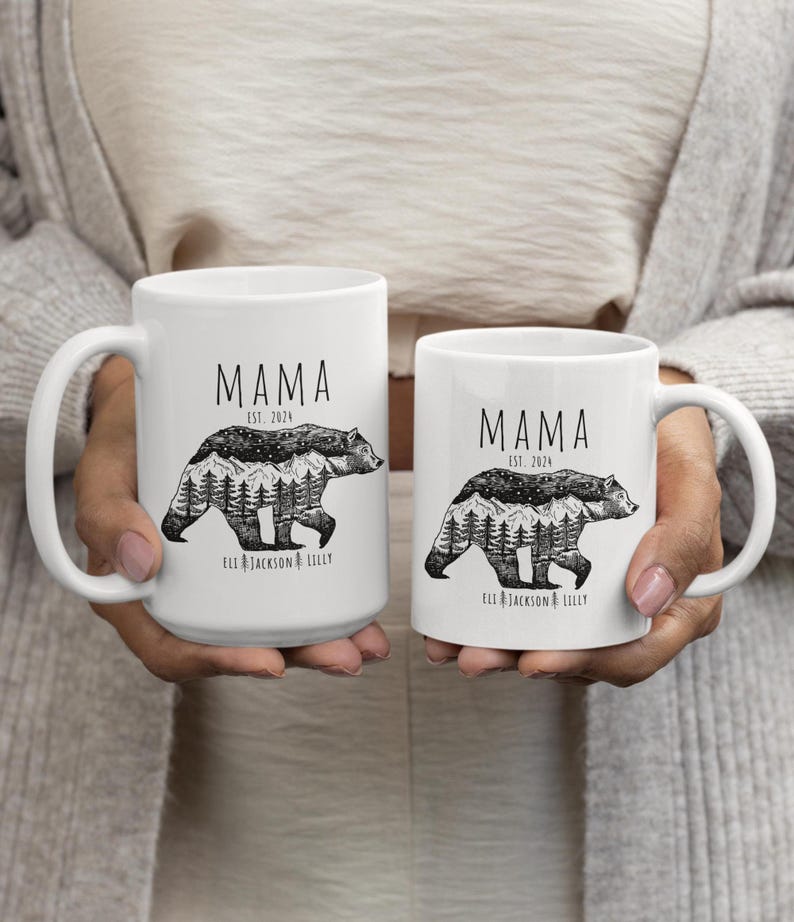 May include: Two white ceramic mugs with black handles. Both mugs feature a black and white illustration of a bear with a mountain range and trees inside its silhouette. The text "MAMA EST. 2024" is printed on both mugs. The text "ELL JACKSON & LILLY" is printed on the bottom of both mugs.