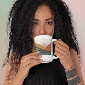 May include: White ceramic mug with a mountain landscape design and the text "Just another" in brown and yellow. The mug is being held by a person with a black tattoo on their wrist.