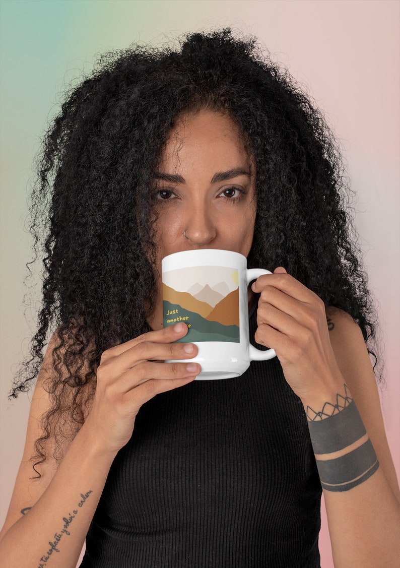 May include: White ceramic mug with a mountain landscape design and the text "Just another" in brown and yellow. The mug is being held by a person with a black tattoo on their wrist.