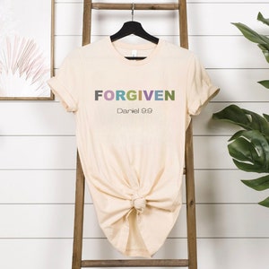 May include: A beige t-shirt with the word "FORGIVEN" in colourful letters and the text "Daniel 9:9" below it.
