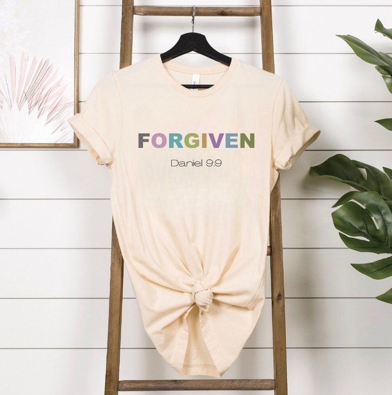 May include: A beige t-shirt with the word "FORGIVEN" in colorful letters and the text "Daniel 9:9" below it.
