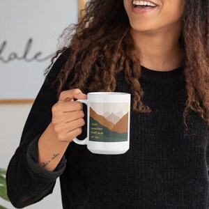 May include: White ceramic mug with a mountain scene and the text "Just another half mile or so..." in yellow. The mug is being held by a person wearing a black sweater and blue jeans.