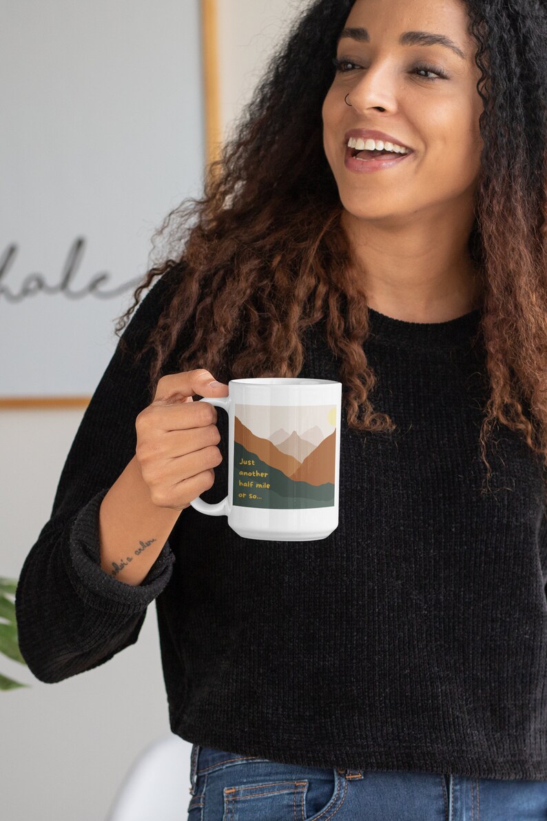 May include: White ceramic mug with a mountain scene and the text "Just another half mile or so..." in yellow. The mug is being held by a person wearing a black sweater and blue jeans.