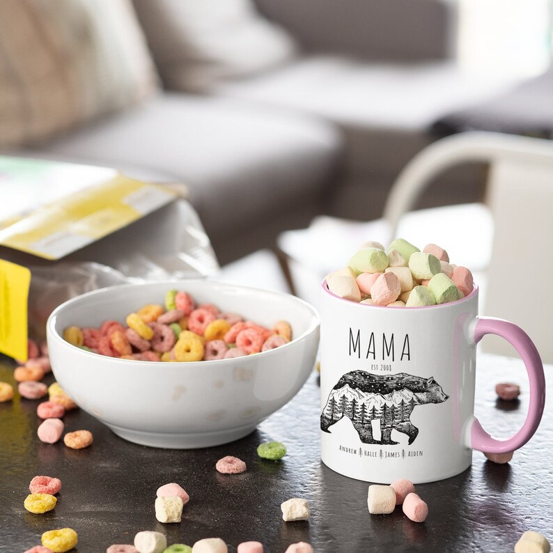 May include: White ceramic mug with a pink rim and handle. The mug has a black and white illustration of a bear with mountains in the background. The text "MAMA EST. 2003" is printed on the mug. The text "ANDREW KAILE JAMES & RIDEN" is printed below the bear.