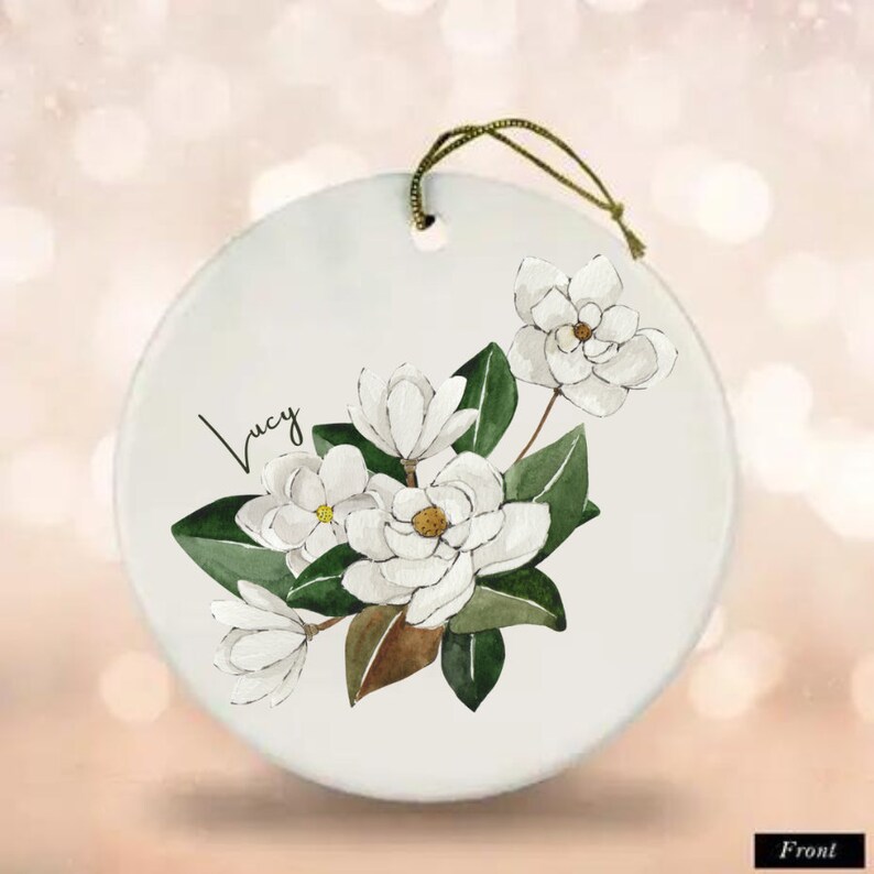 Może przedstawiać: Biała ozdoba ceramiczna z akwarelowym wzorem białych kwiat&oacute;w magnolii i zielonych liści. Ozdoba ma sznurek do zawieszenia i imię "Lucy" napisane kursywą z przodu.