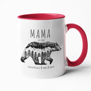 May include: White ceramic mug with a red rim and handle. The mug has a black and white illustration of a bear with a mountain range and trees inside its silhouette. The text "MAMA EST 2000" is above the bear and the text "ANDREW HALLE JAMES AIDEN" is below the bear.