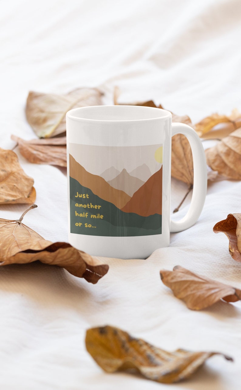 May include: White ceramic mug with a graphic of a mountain range with a sun in the sky. The text on the mug reads "Just another half mile or so..."