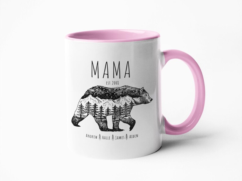 May include: White ceramic mug with a pink rim and handle. The mug features a black and white illustration of a bear with a mountain range and stars inside. The text "Mama Est 2003" is above the bear and the text "Andrew Halle James Aiden" is below the bear.