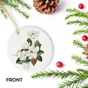 Może przedstawiać: Biała ceramiczna ozdoba z kwiatowym wzorem przedstawiającym białe kwiaty magnolii i zielone liście. Ozdoba jest spersonalizowana imieniem "Lucy".