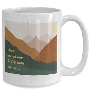 May include: White ceramic coffee mug with a mountain landscape design. The mug has a handle and features the text "Just another half mile or so..." in yellow.