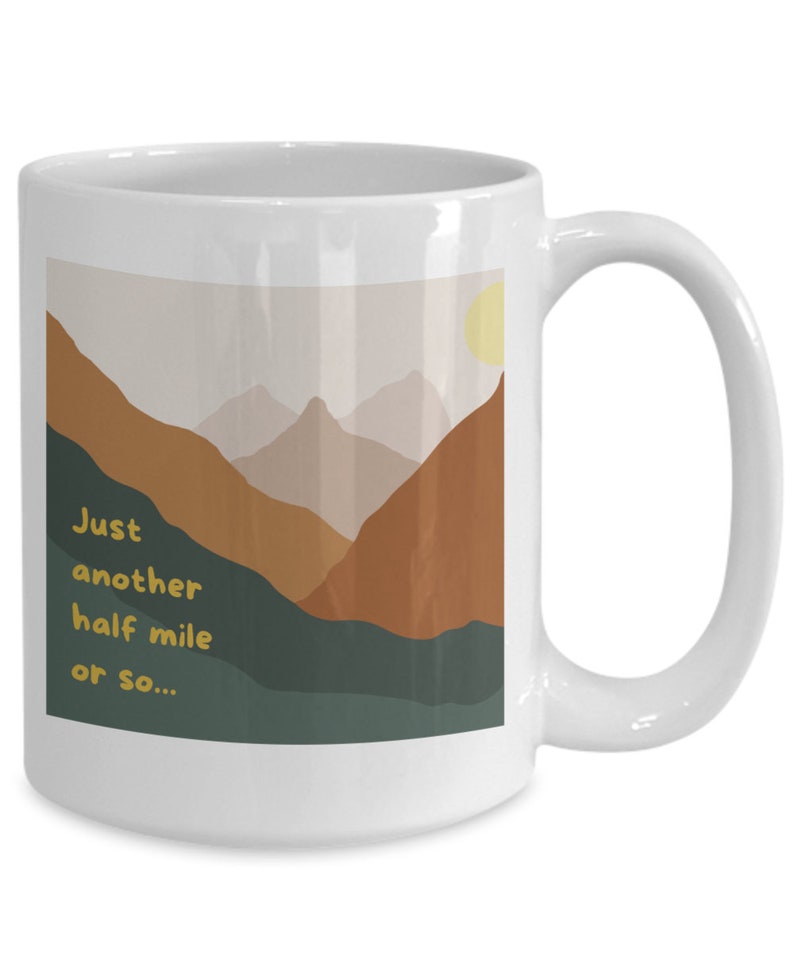 May include: White ceramic coffee mug with a mountain landscape design. The mug has a handle and features the text "Just another half mile or so..." in yellow.