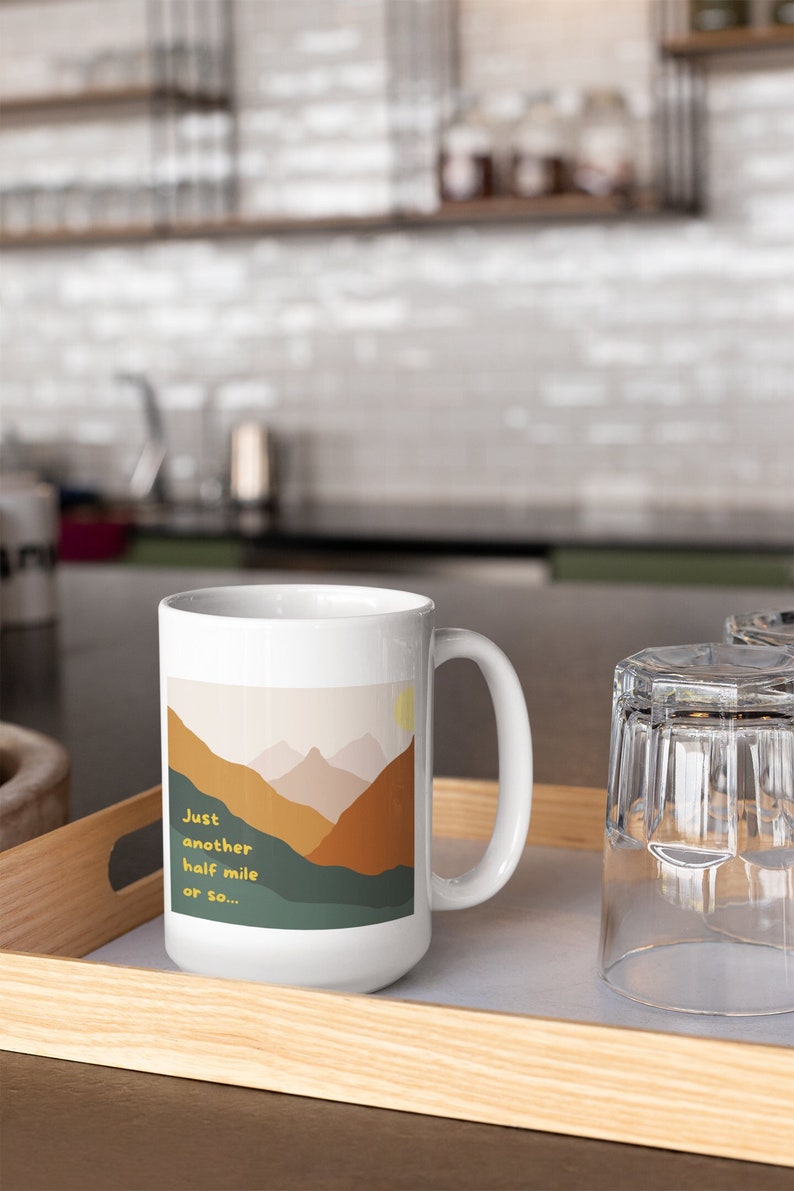 May include: White ceramic mug with a mountain landscape design. The mug has a handle and says "Just another half mile or so..." in yellow text.