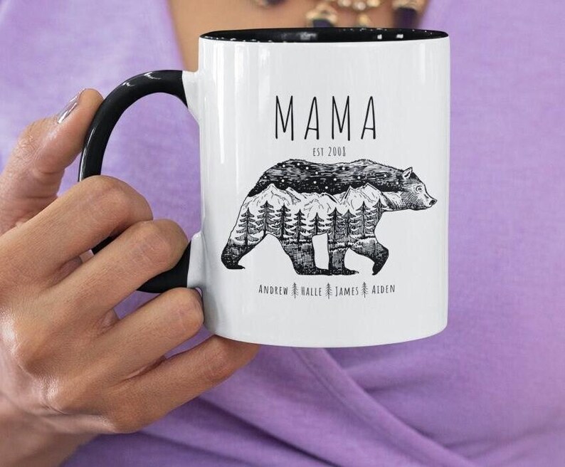 May include: White ceramic mug with a black handle. The mug has a black rim and says "MAMA EST 2001" with a black and white illustration of a bear with mountains and trees inside. The text "ANDREW & HALLE & JAMES & AIDEN" is printed below the bear.