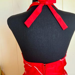 May include: A red halter dress with a white floral pattern and a zipper closure. The dress features a red bow at the neck and is displayed on a black mannequin.