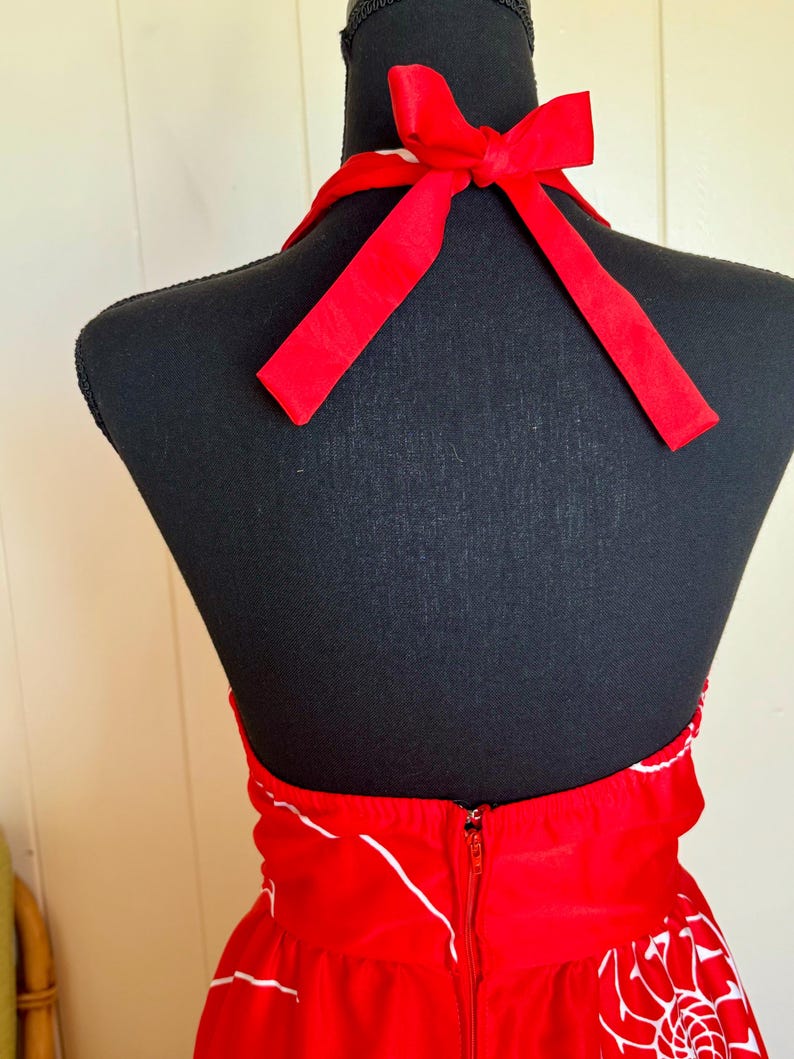 May include: A red halter dress with a white floral pattern and a zipper closure. The dress features a red bow at the neck and is displayed on a black mannequin.