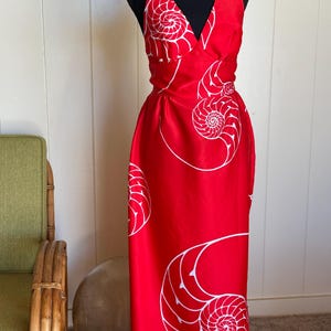 May include: A red halter dress with a white nautilus shell pattern. The dress has a V-neck and a flowing, floor-length skirt. The dress is displayed on a black mannequin.