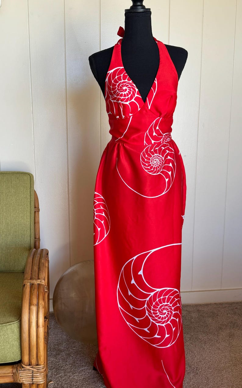 May include: A red halter dress with a white nautilus shell pattern. The dress has a V-neck and a flowing, floor-length skirt. The dress is displayed on a black mannequin.