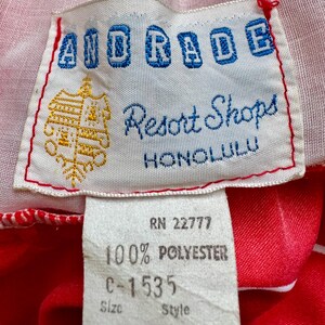May include: Close-up of a vintage clothing tag. The tag reads "AIDRADE Resort Shops HONOLULU" in blue and white lettering. Below, a smaller tag indicates "100% POLYESTER" and size 8. The background is red and white fabric.