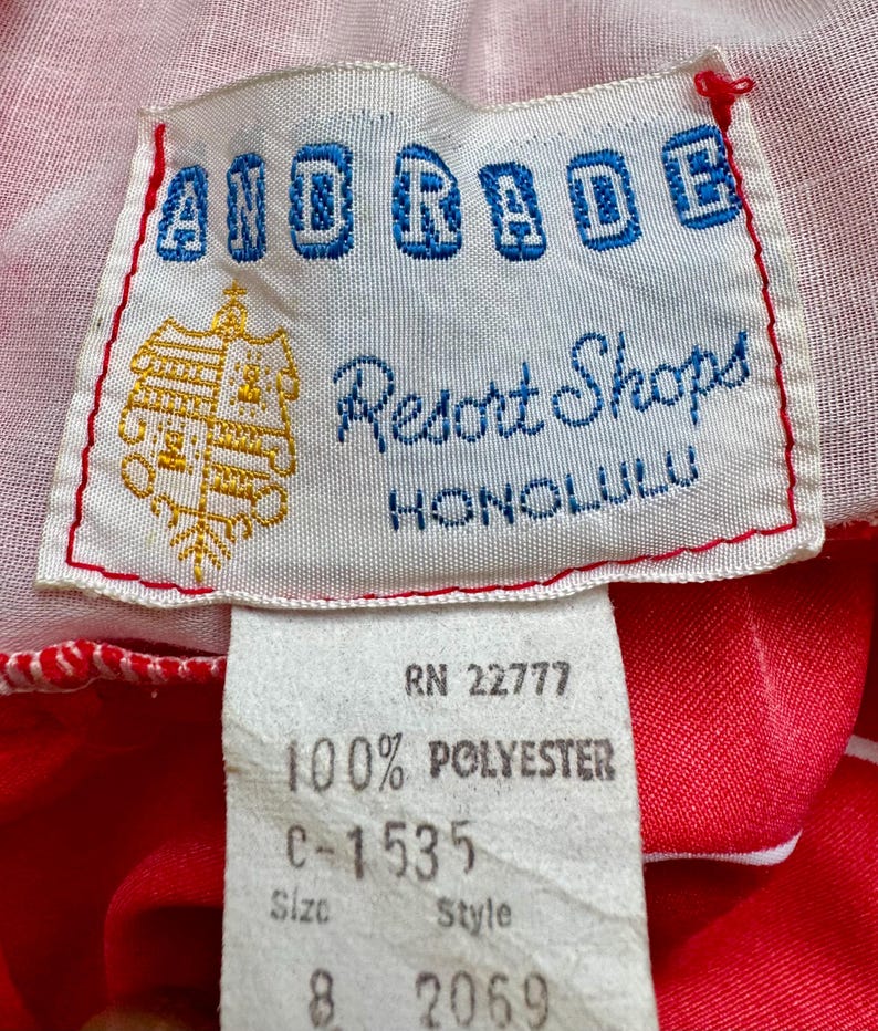 May include: Close-up of a vintage clothing tag. The tag reads "AIDRADE Resort Shops HONOLULU" in blue and white lettering. Below, a smaller tag indicates "100% POLYESTER" and size 8. The background is red and white fabric.