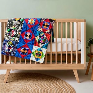 May include: A colorful patchwork quilt draped over a light wood crib. The quilt features squares of red, blue, green, and black fabric with geometric patterns. The crib has a white mattress and is set on a round jute rug.