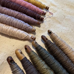 Peut inclure: Une collection de fuseaux en bois enroulés de fils de différentes teintes, notamment crème, beige, marron, vert olive et rose poudré. Chaque fuseau présente un petit trou à son extrémité, avec le fil enroulé étroitement autour du corps en bois.