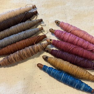 May include: A collection of twelve wooden bobbins wrapped in various colors of yarn, including shades of beige, brown, gray, pink, red, purple, yellow, and blue. Each bobbin features a tapered shape with a small hole at one end for thread.