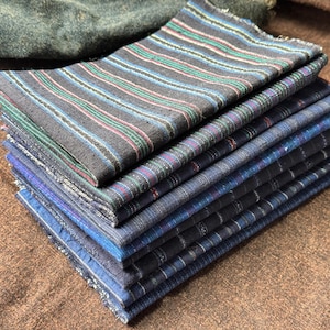 Può includere: Una pila di quadrati di tessuto piegati in varie tonalità di blu e grigio. Il quadrato superiore presenta un motivo a righe con accenti turchese, rosa e verdi. Il tessuto sembra essere tessuto, con una superficie leggermente strutturata. I quadrati sono impilati su una superficie marrone.