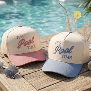 May include: Two baseball caps with cream-colored crowns and colored brims, one pink and one blue. Both caps feature the embroidered text 'IT'S Pool TIME' on the front. A pair of sunglasses and two glasses with lime slices are also present.