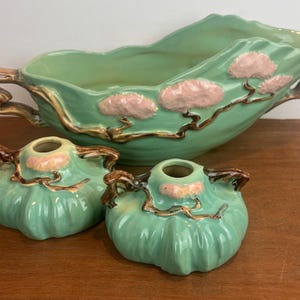 May include: A set of three ceramic pieces: a large, leaf-shaped bowl and two small, round vases. The pieces are a light green color with pink and brown accents. The bowl has handles and decorative pink cloud-like shapes.