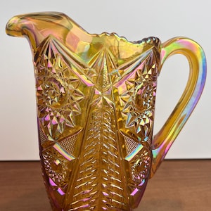 Op de afbeelding: Een iriserende amberkleurige glazen kan met een sterren- en geometrisch patroon. De kan heeft een handvat en een tuit om uit te schenken. Het glas reflecteert tinten goud, paars en groen. De kan is waarschijnlijk vintage of antiek.