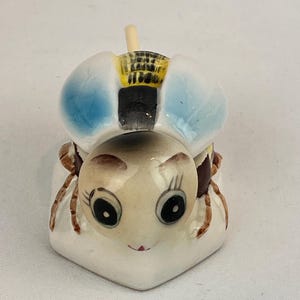 May include: A ceramic bee figurine with a smiling face, large black eyes, and blue wings. The bee has brown legs and a yellow and black striped back. The figurine is on a white background.