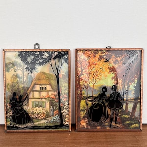 May include: Two framed vintage silhouette art pieces. One depicts a couple dancing in front of a cottage, the other shows a couple strolling in a park. Both have copper-colored frames and are set against a colorful background.