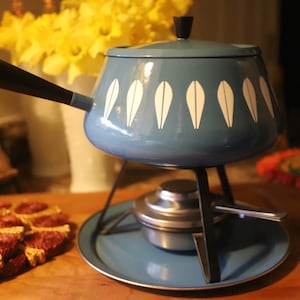 May include: A vintage fondue pot in a teal blue color with white leaf-shaped designs. It has a black handle and sits on a matching stand with a burner. The pot is a classic piece for entertaining.