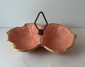 Può includere: Un piatto da portata in ceramica rosa con un bordo dorato e un motivo a foglie. Il piatto ha un manico al centro ed è diviso in due sezioni.