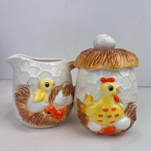 May include: A white ceramic creamer and sugar bowl set. The creamer has a handle and a chick in a nest design. The sugar bowl has a lid with a white egg-shaped knob and a chick design. Both pieces have a chicken wire pattern.