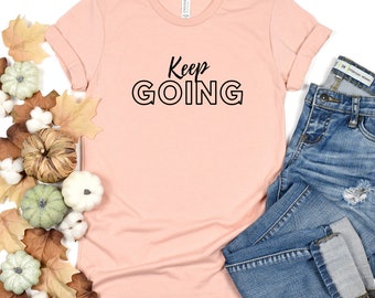 May include: A peach-colored t-shirt with the text "Keep GOING" in black outlined letters. The shirt is shown on a white background with a pair of blue jeans.