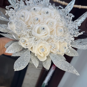 Pode incluir: Um buquê de noiva com rosas brancas e folhas de glitter prata. O buquê é decorado com cristais transparentes.