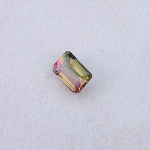 May include: A rectangular gemstone with a gradient of colours, including pink, yellow, and green. The gemstone is set against a white background. The cut of the stone is a rectangle.