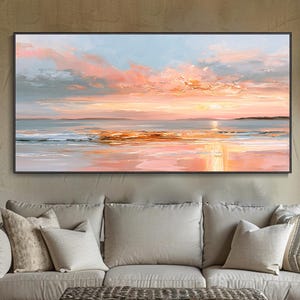 Op de afbeelding: Een rechthoekig landschapsschilderij met een zonsondergang boven een kalm wateroppervlak, met zachte roze, oranje en blauwe tinten in de lucht en reflecties op het water, ingelijst in een dunne zwarte lijst boven een bank met kussens.