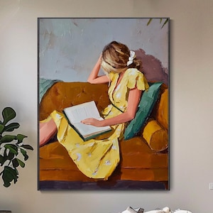 Pode incluir: Uma pintura a óleo retangular sobre tela retratando uma pessoa de cabelos loiros vestindo um vestido amarelo, sentada em um sofá cor de mostarda enquanto lê um livro aberto. A obra apresenta uma almofada azul-petróleo e um fundo de tons suaves.