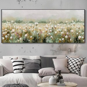 May include: A rectangular canvas art piece depicting a field of white flowers under a soft, muted sky. The painting has a neutral color palette with touches of green and pink. The artwork is framed in a black border and hangs above a light-colored sofa.