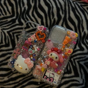 May include: Two clear phone cases decorated with various charms, including Hello Kitty and My Melody characters, hearts, and beads. One case has a "HUG ME" heart charm. The other has a "BE MINE" heart charm. The background is a black and white zebra print.