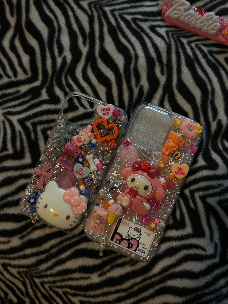 May include: Two clear phone cases decorated with various charms, including Hello Kitty and My Melody characters, hearts, and beads. One case has a "HUG ME" heart charm. The other has a "BE MINE" heart charm. The background is a black and white zebra print.