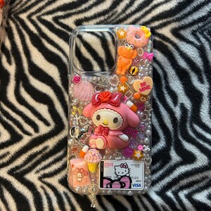 May include: Clear phone case decorated with a pink My Melody figure, a heart that says "Be Mine", and various charms. The case also features a Hello Kitty credit card design and is embellished with pearls and other decorative elements.